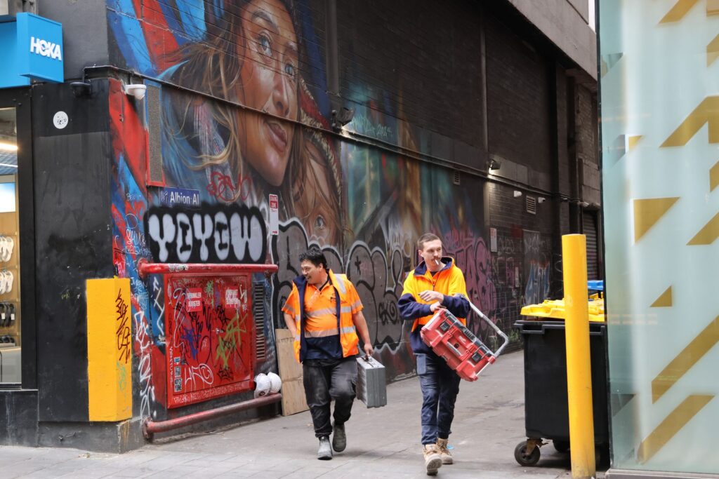 Two workers in orange safety uniforms walking through a graffiti-covered alley in Melbourne, with a large mural of a woman on the wall and a red utility box covered in tags.