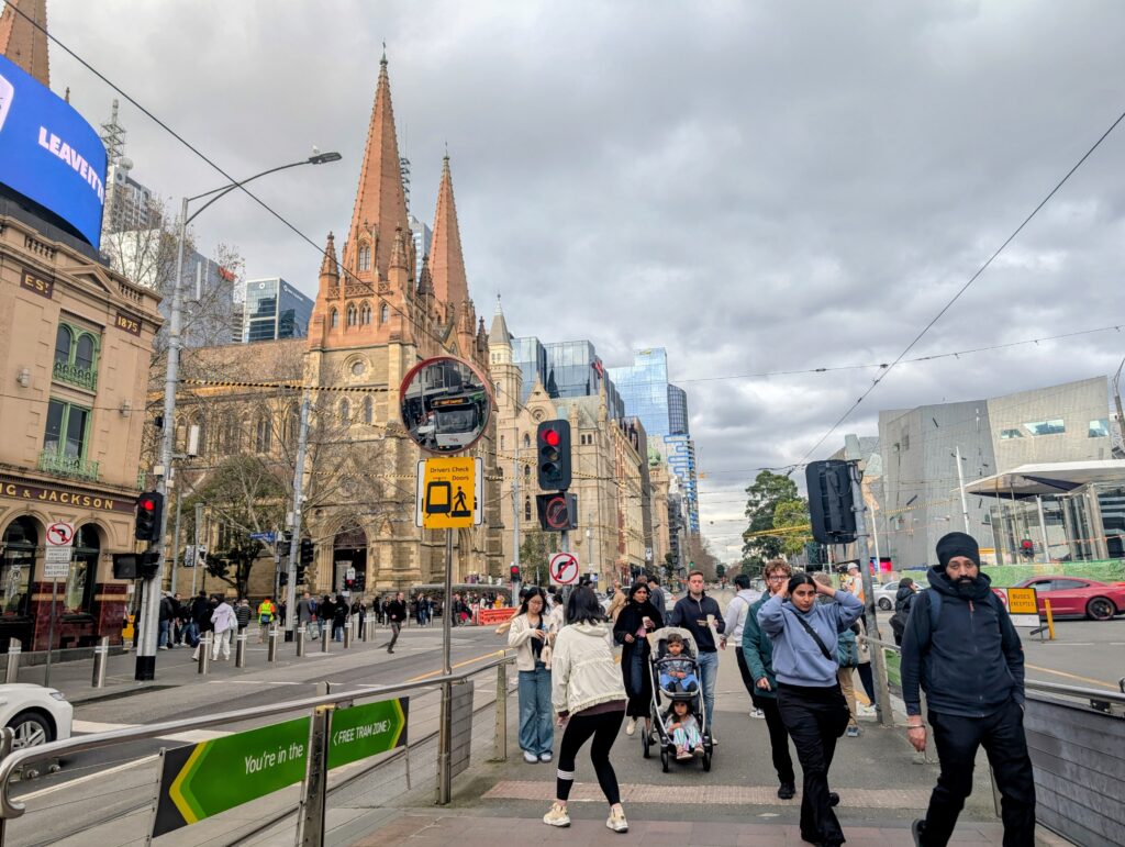 Pedestrians crossing near St Paul’s Cathedral in Melbourne on a cloudy winter day, with trams, traffic lights, and city buildings in the background.